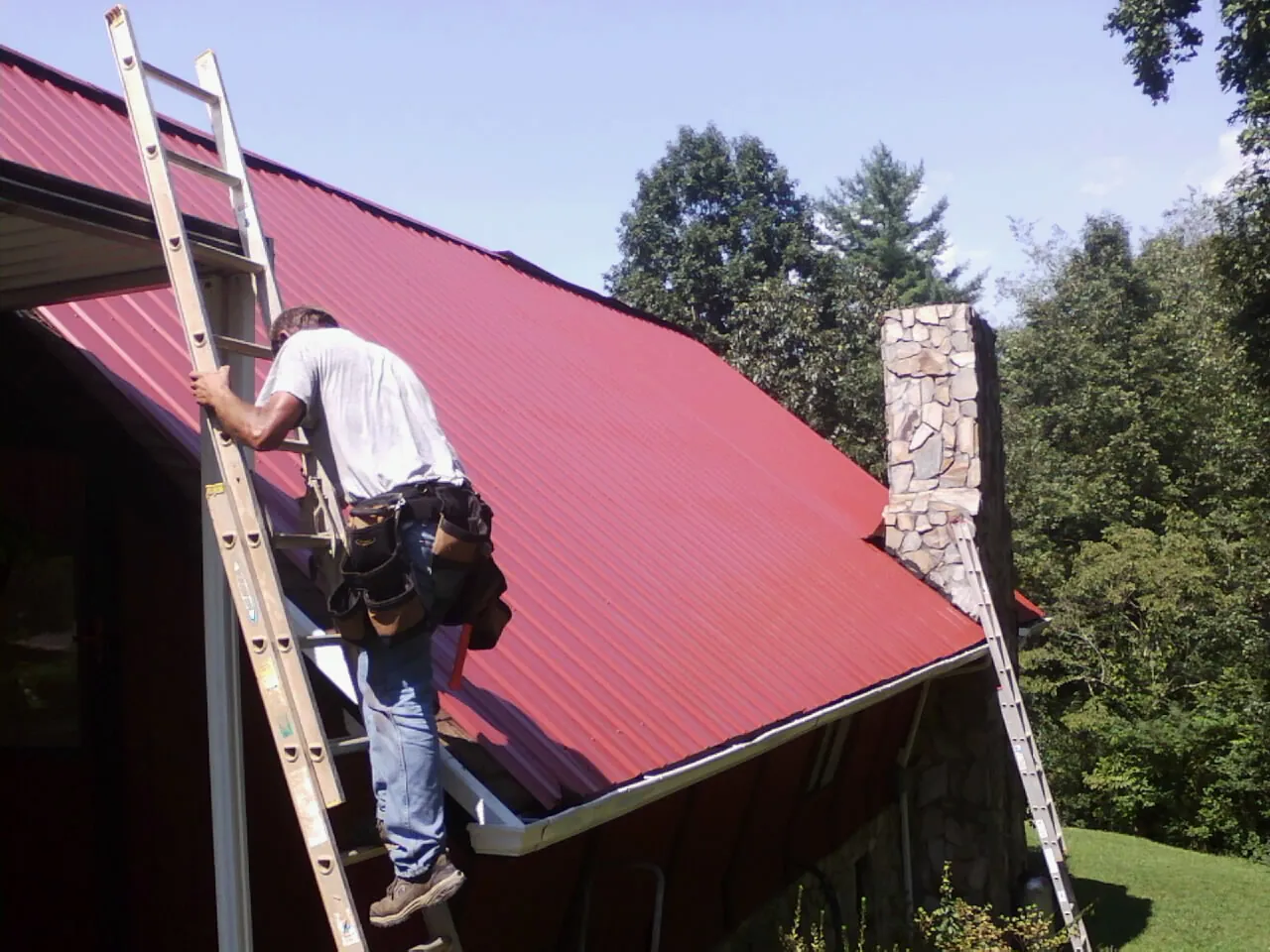 Roofer on a ladder beside a red metal roof and stone chimney — Soffit & Fascia Repair by Roofing Brothers in Barnes City